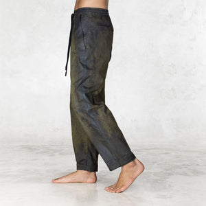 Hand painted half olive half black pants worn by a person shown feet to waist facing the side on a light gray background