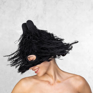 Woman from the chest up wearing a fuzzy black balaclava with horns that is covering half her face. She is spinning her head so the fur is in motion.