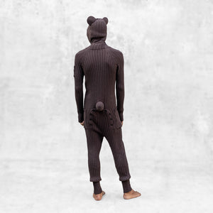 A person wearing a brown hooded knit bear onesie with a round tail standing against a light gray background