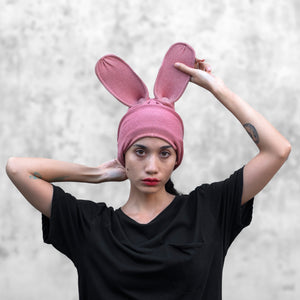 A person wearing a pink bunny balaclava with visible ears and a black top against a grey background.
