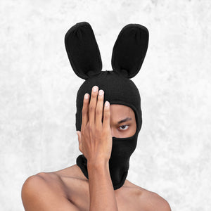 Person wearing a black bunny balaclava that covers their mouth and is covering one eye with their hand in front of a light gray background