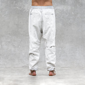 A man standing barefoot and shirtless from the feet to the waist facing back wearing a pair of white parachute style linen pants. 