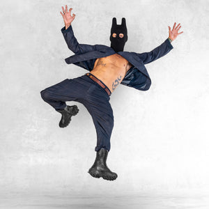 A man in an Indigo jacket un buttoned to the top and Indigo cargo style pants with a balaclava on and jumping in a surprise position. 
