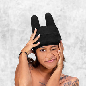 Person wearing a cotton knit black beanie with rounded ears with their hands on their face against a light gray background