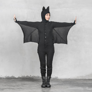 A person wearing a black bat onesie with arms spread out resembling bat wings standing against a gray textured background