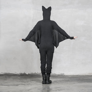 A person wearing a black bat onesie with arms spread out in a V-shape, resembling bat wings standing against a gray textured background