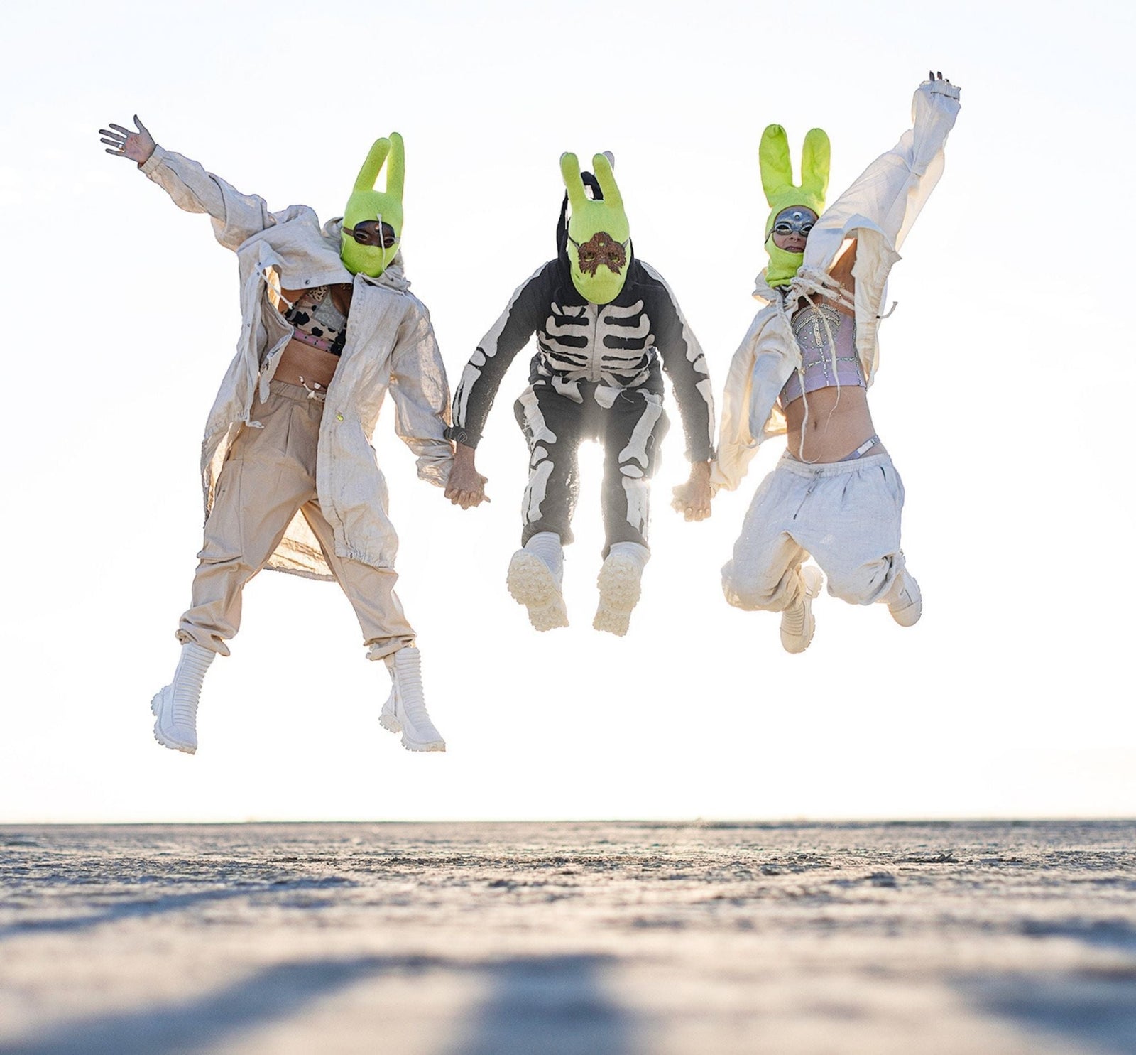 Three people in bold desert fashion and neon bunny masks jumping mid-air at sunrise on the playa during Burning Man