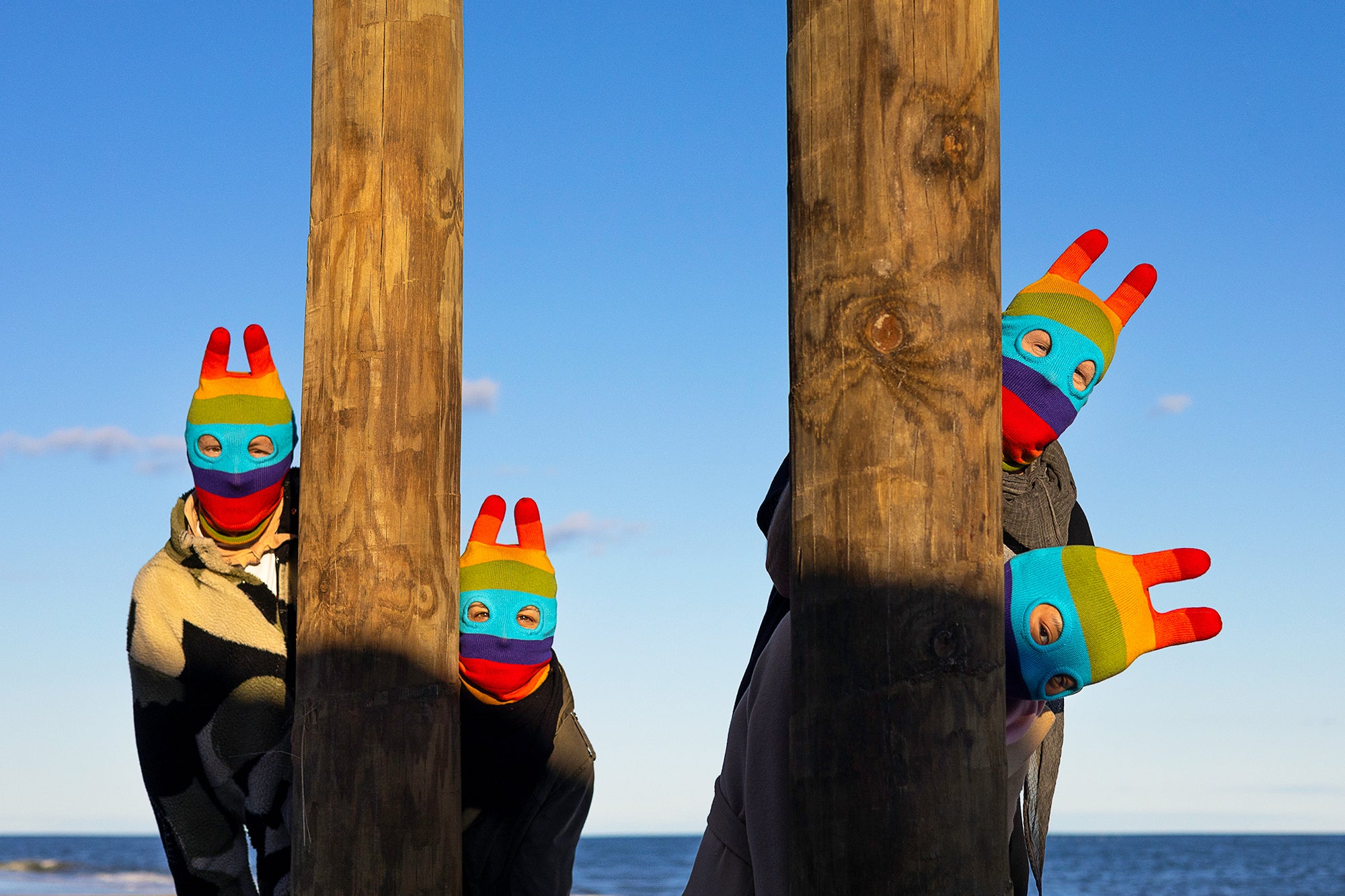 a group of 4 people in rainbow balaclavas peeking out from behind wooden pillars