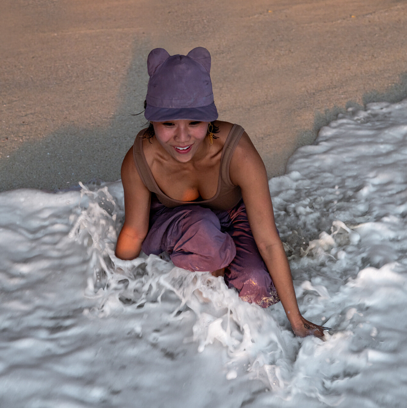 Person wearing a purple hat and pants sitting in shallow water on a beach.