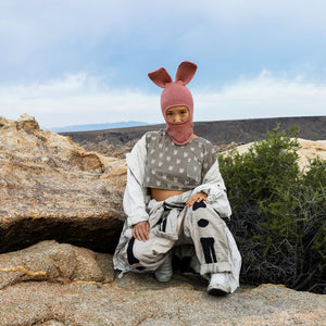 Person in a pink bunny balaclava mask and beige clothing kneeling down on rock with nature in the background