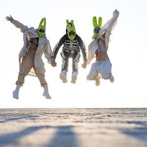 Three people holding hands and jumping in the air wearing green balaclava masks on a sandy landscape with the sun behind them