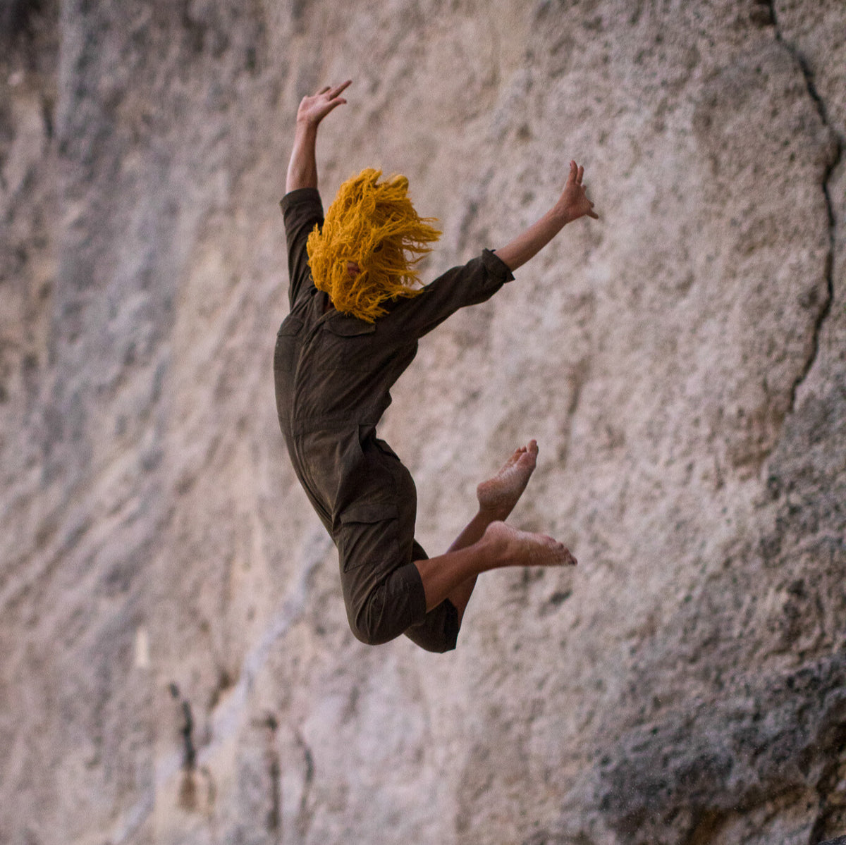 Person with yellow hair in a black outfit leaping against a rocky cliff.