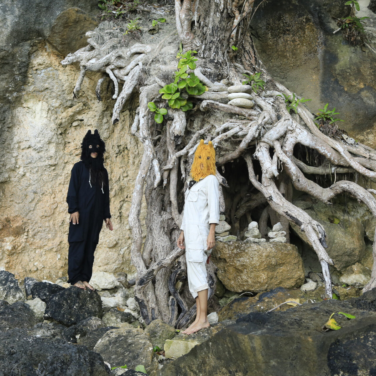 Two people in animal costumes standing near a cave entrance with roots and rocks.