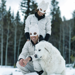 Two people in bear masks sitting next to a large white dog in a snowy forest.