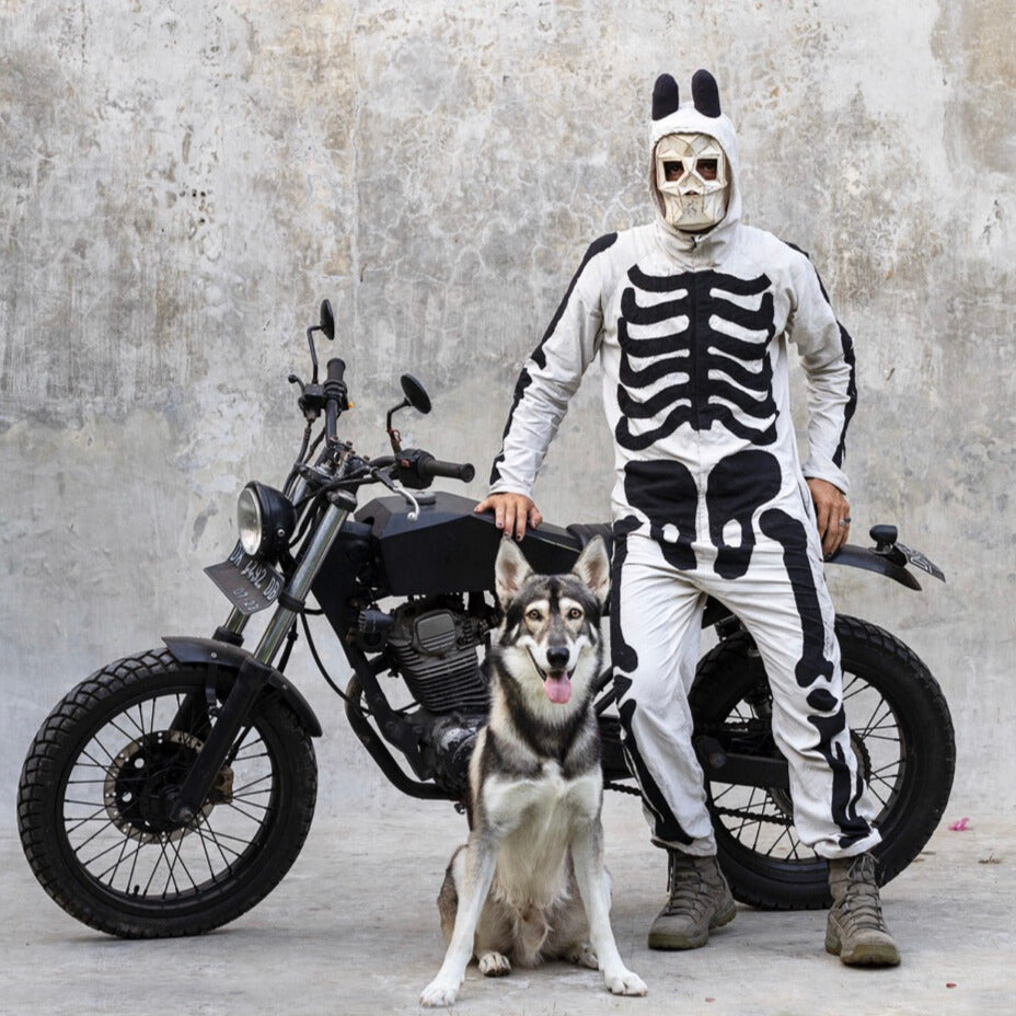 Person in skeleton costume with a dog on a motorcycle against a textured wall.
