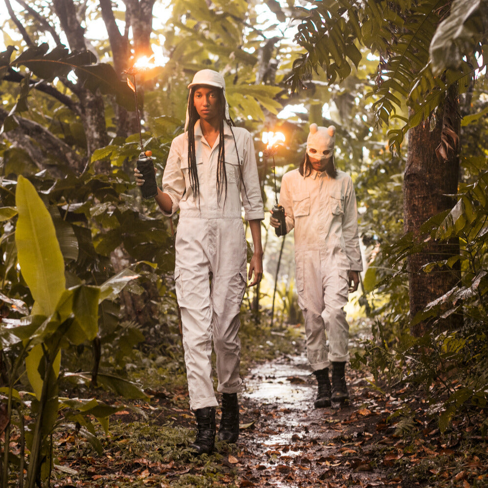 Two people wearing white jumpsuits and masks and black leather boots walking through a forest in the rain and holding torches