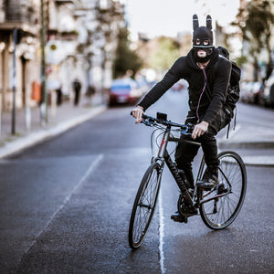 Person riding a bicycle on a city street wearing a dark outfit and bunny ears mask.