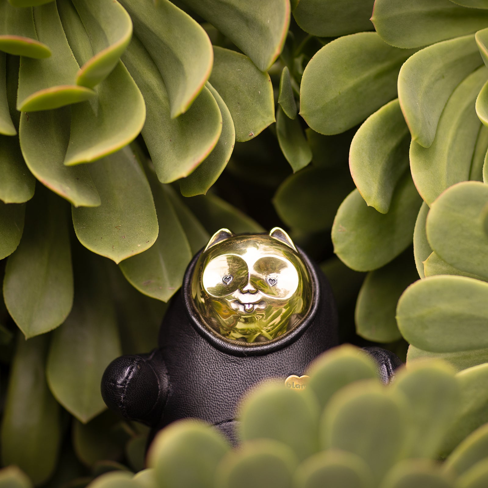 An animal figurine resembling a cat with a brass face with its tongue sticking out and a brass body surrounded by green succulents 