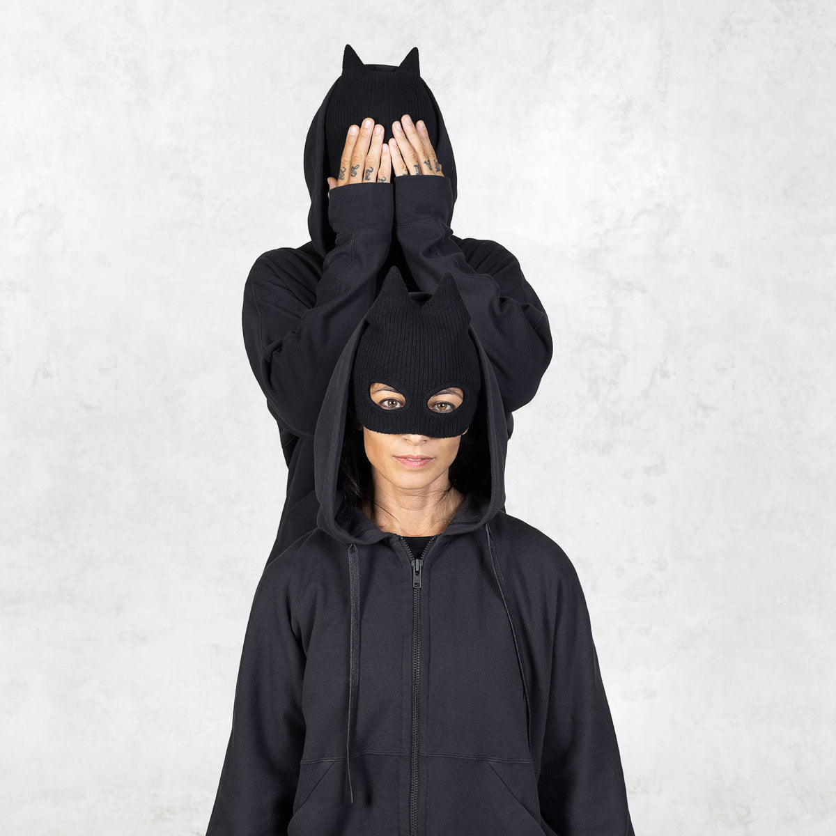 Two people wearing matching black hoodies and black masks with spiked ears against a light gray background.