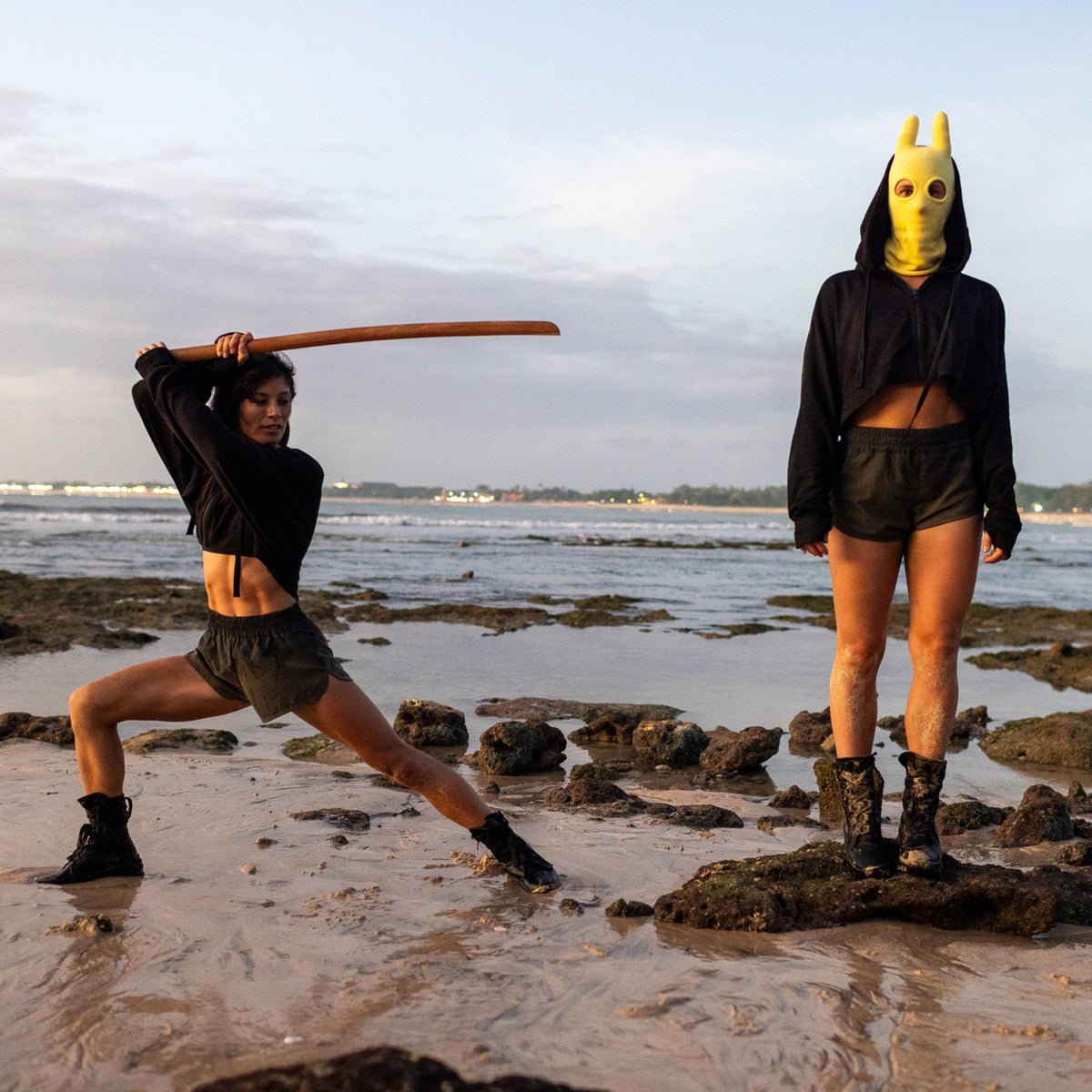 Two people on a beach, one with a bokken sword and the other wearing a mask, with ocean and sky in the background.