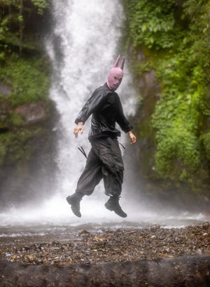 Person in oversized olive pants and zip up hooded jacket with a pink sike balaclava in front of a waterfall. 