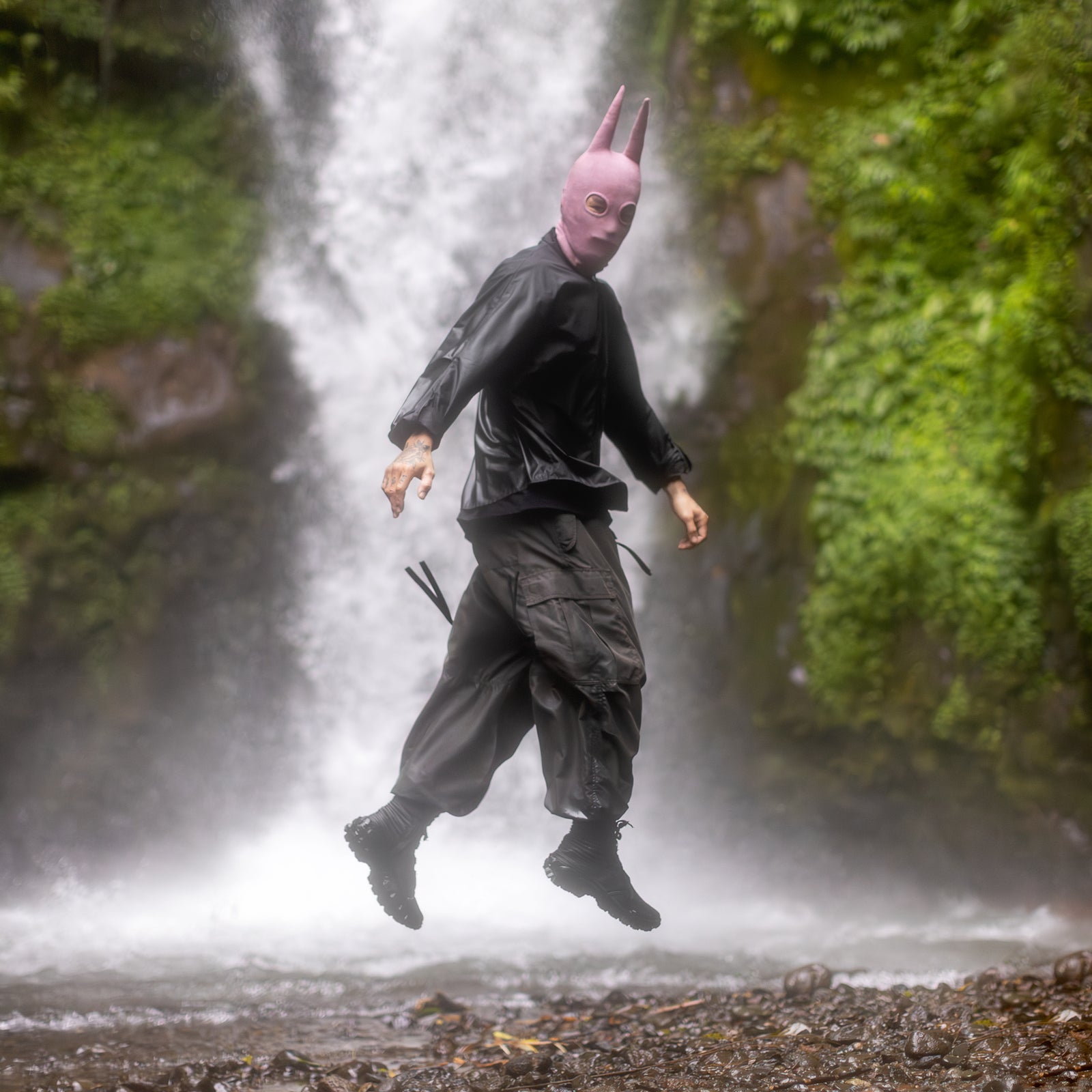 Person in a black shirt, olive green pants, black boots, and a pink balaclava mask with tall spikey ears jumping in front of a waterfall in a natural setting