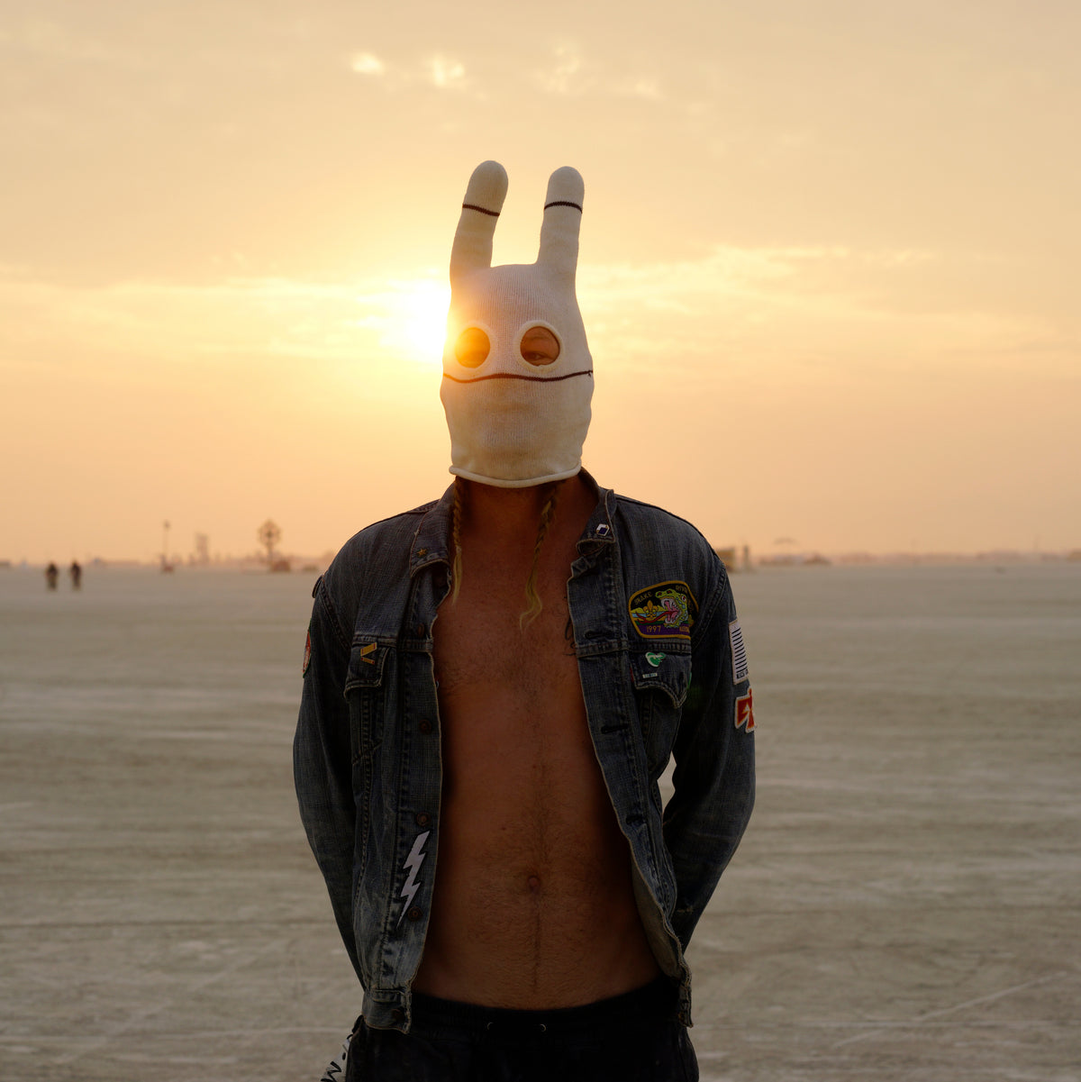 A person wearing a white mask with ears and a denim jacket standing in the desert with the sunset behind them 