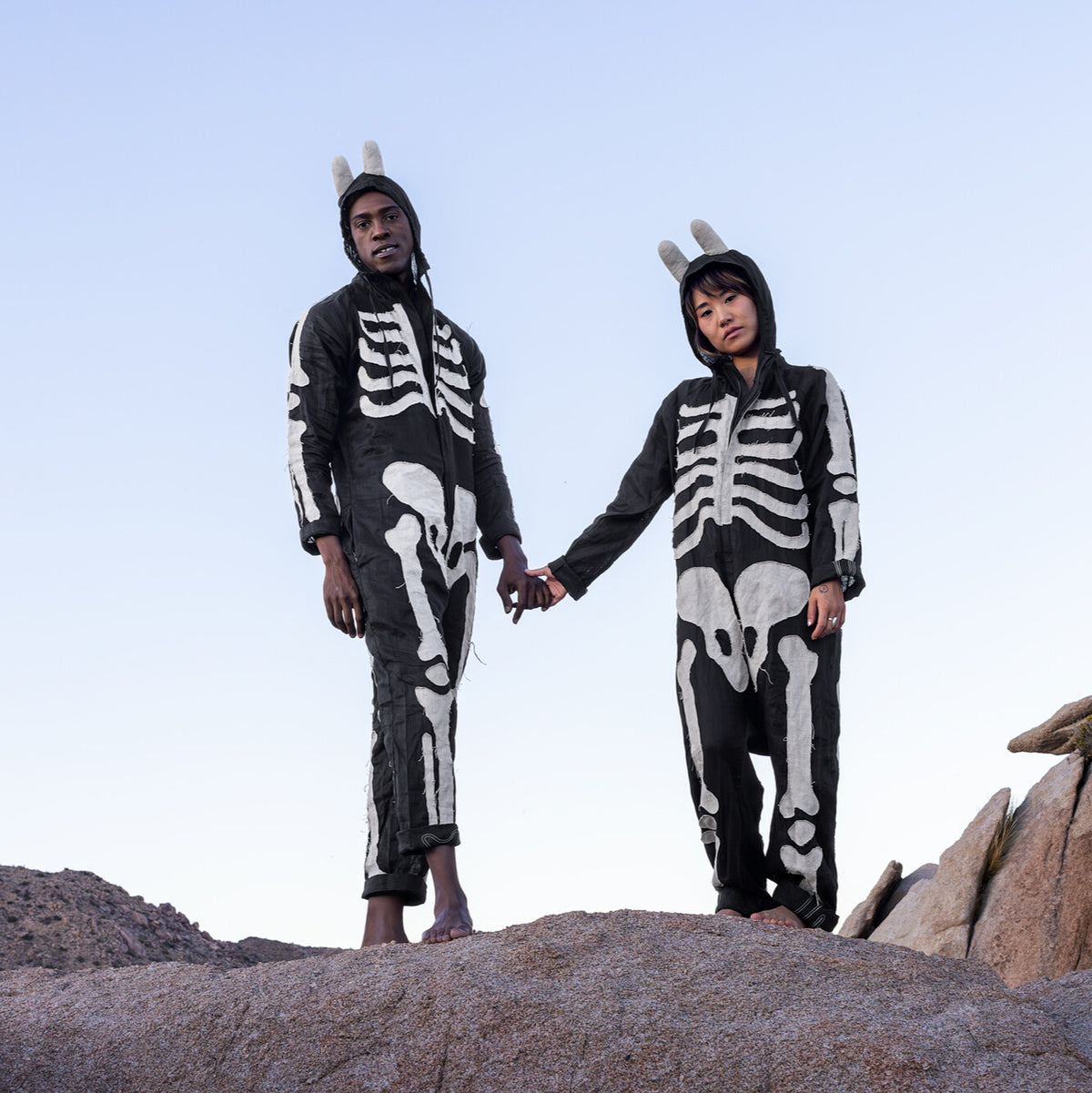 Two people wearing skeleton-themed outfits standing on rocky terrain with a clear sky.