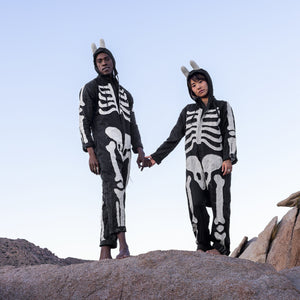 Two people wearing skeleton-themed outfits standing on rocky terrain with a clear sky.
