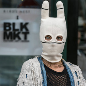 Person wearing a white bunny mask with a blurred background featuring 'BLK MKT' sign.