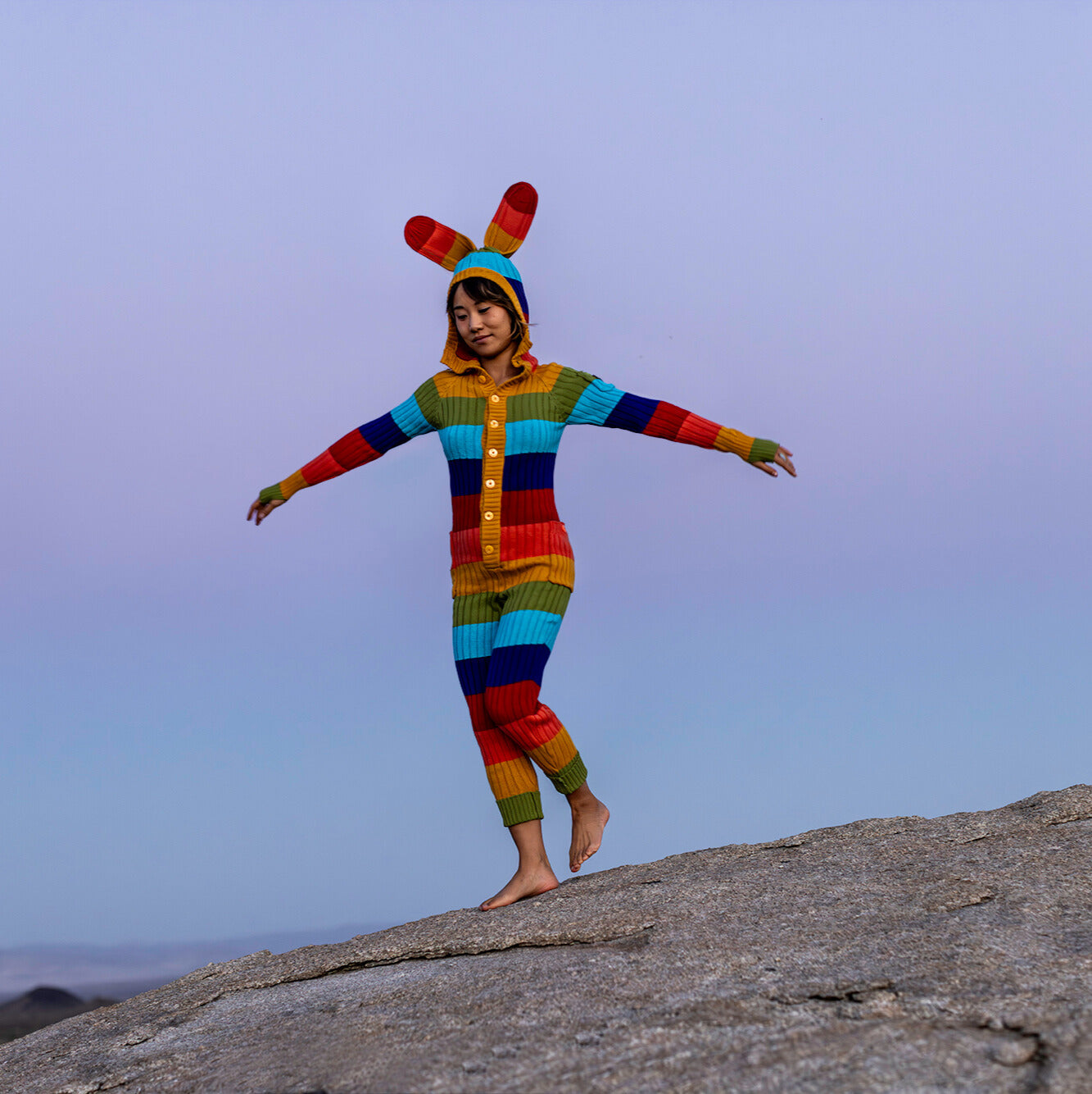 Person wearing a colorful knitted bunny costume standing on a rock with a clear sky background