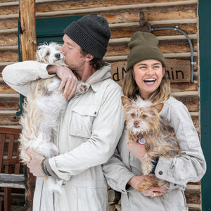 Two people wearing white jumpsuits and beanies holding their dogs in front of a log cabin