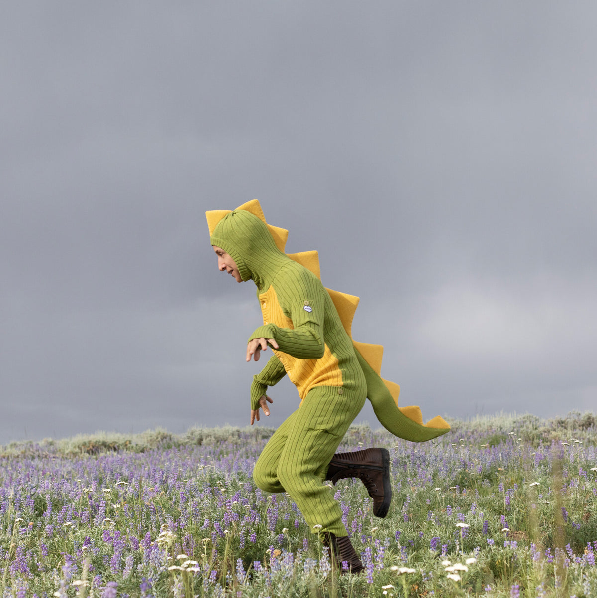 Person wearing a green dinosaur costume running through a field with purple flowers and a gray sky.