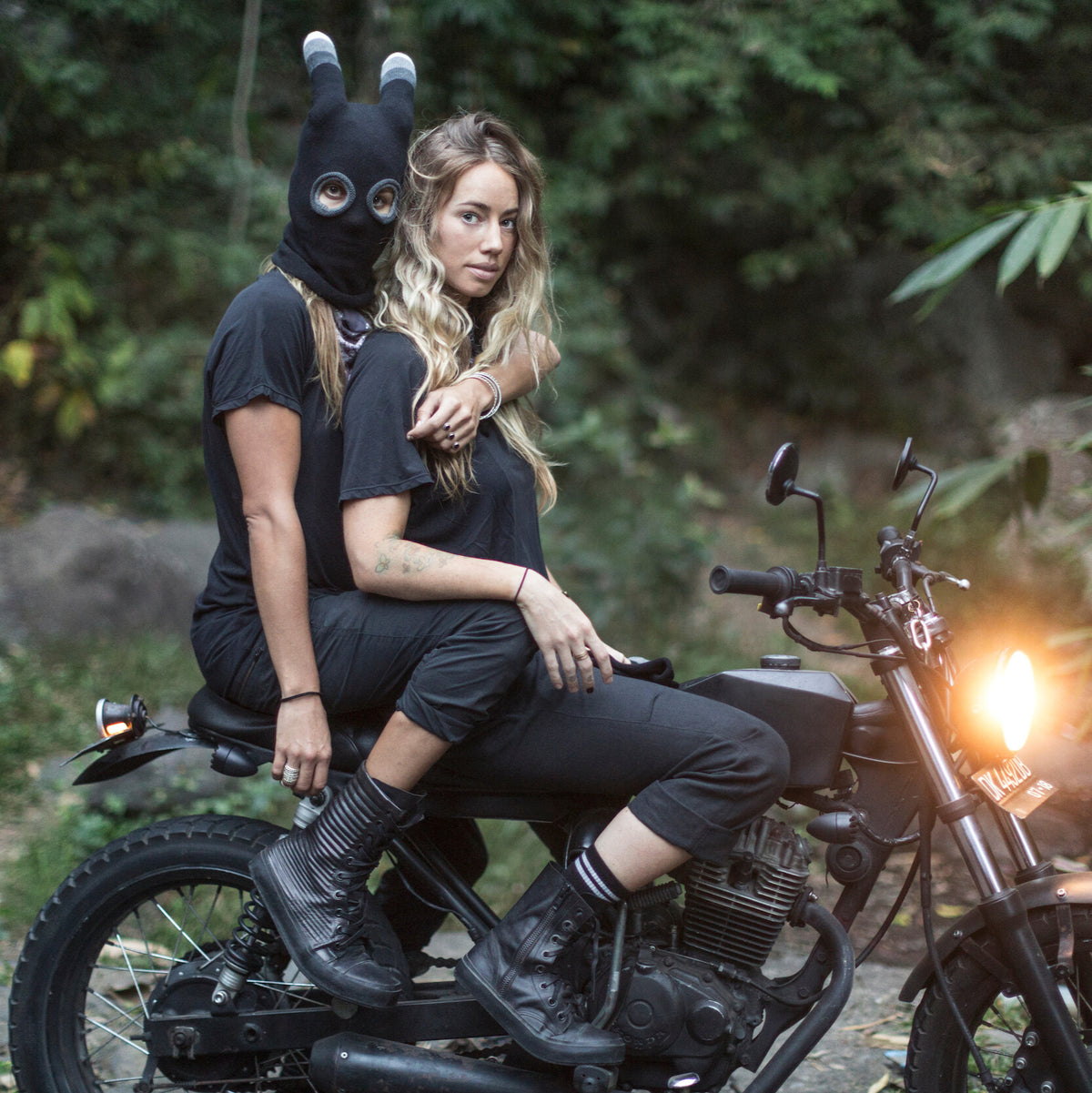 Person wearing a black mask sitting on a motorcycle in a forest setting