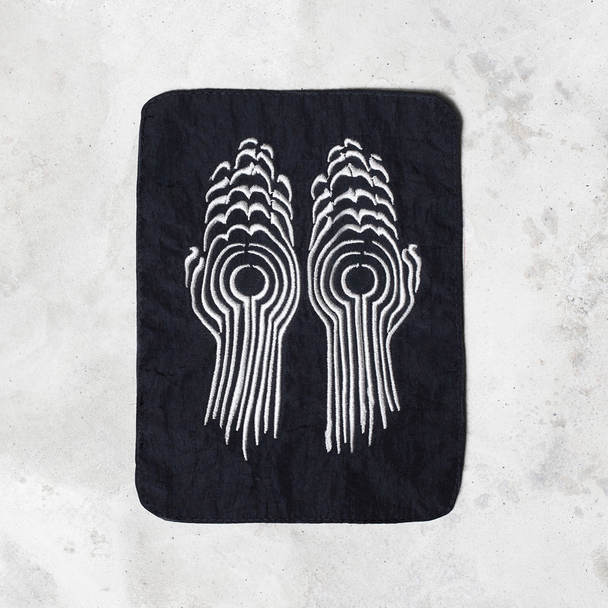 A rectangular black patch with rounded corners featuring a white embroidered design of two stylized hands facing each other, laid flat on a light gray textured surface.