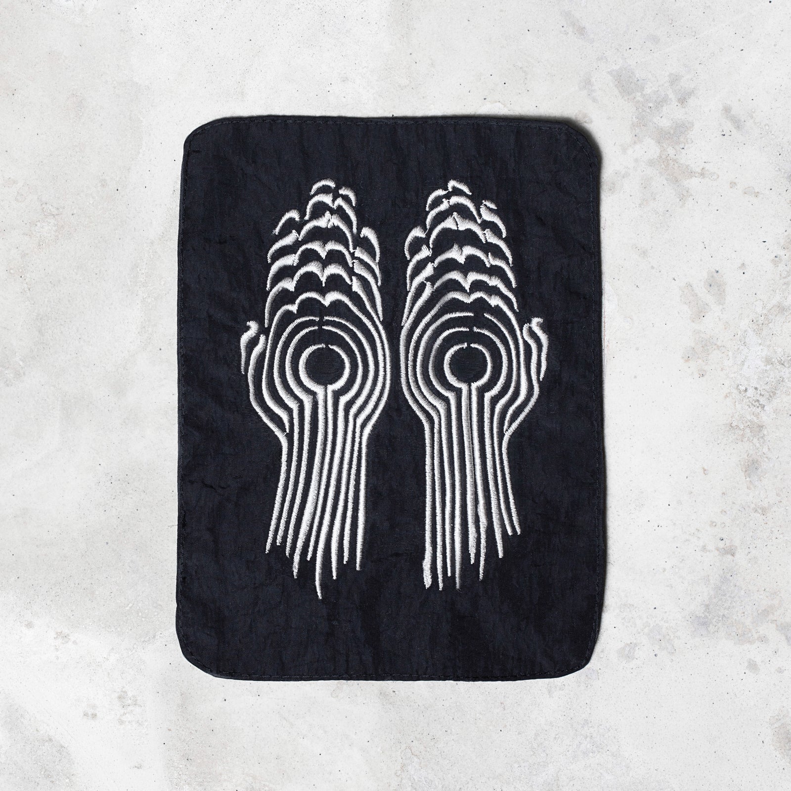 A rectangular black patch with rounded corners featuring a white embroidered design of two stylized hands facing each other, laid flat on a light gray textured surface.