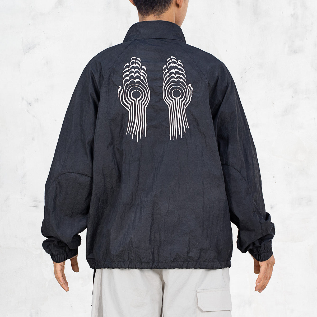 Person wearing a black nylon jacket with a while line embroidered design on the back standing in front of a light gray background