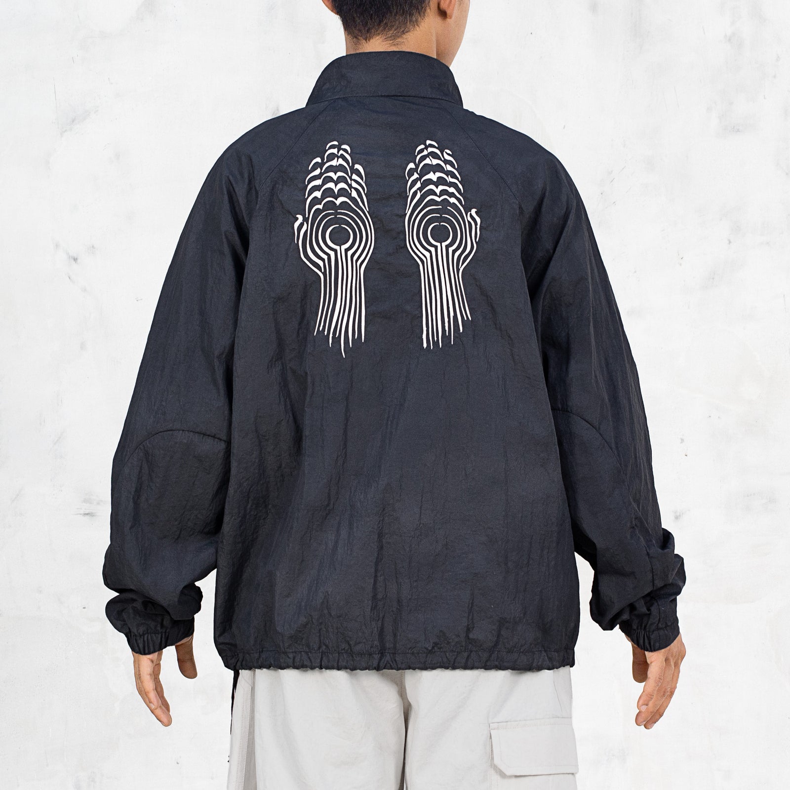 Person wearing a black nylon jacket with a while line embroidered design on the back standing in front of a light gray background