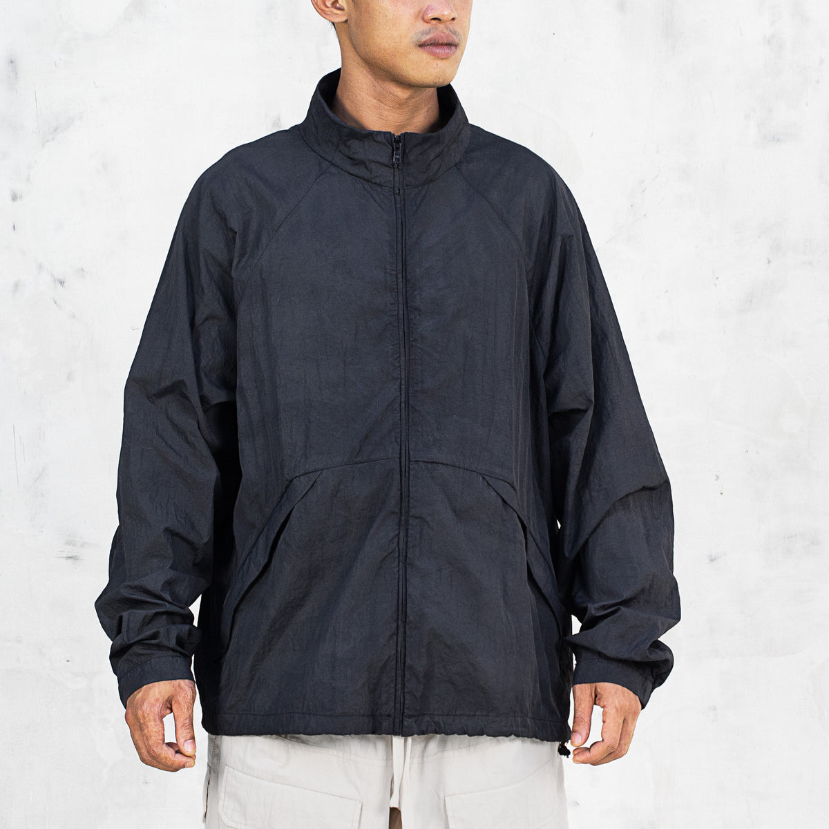 Person wearing a black zip up nylon mock neck jacket with sloped pockets standing in front of a light gray background 