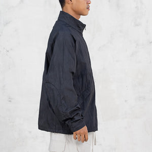 Side profile of a person wearing a black lightweight nylon jacket and light pants against a light gray background