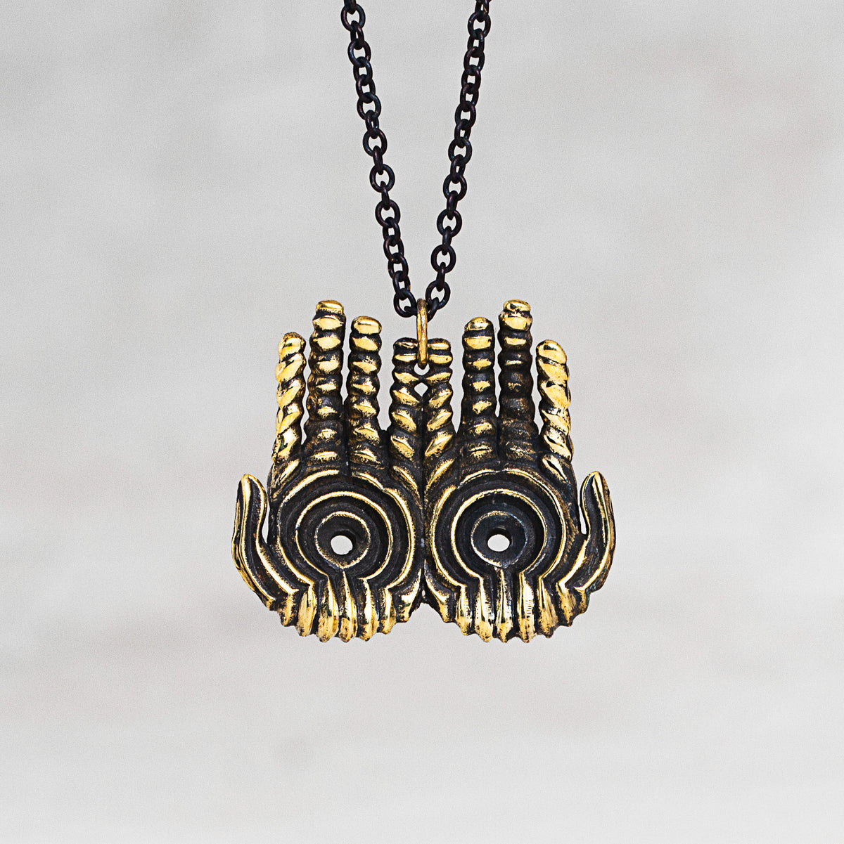 A close-up of a necklace featuring a double hand pendant with a textured gold finish and black enamel details, suspended on a black chain against a light gray background.