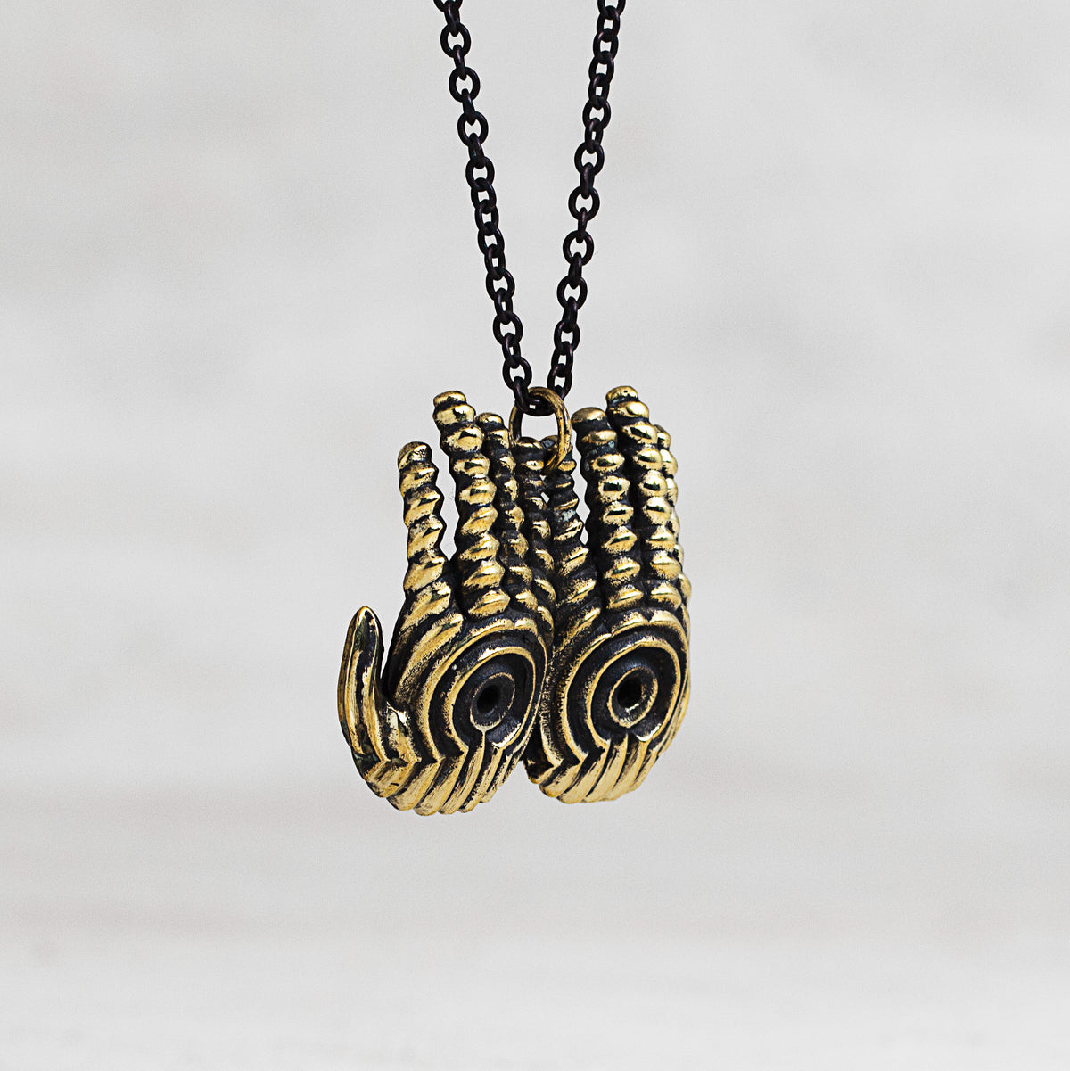 A close-up of a necklace featuring a textured, double-hands pendant in an antique gold finish with black recessed detailing, suspended from a black chain against a light gray background.