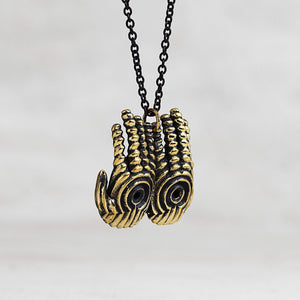 A close-up of a necklace featuring a textured, double-hands pendant in an antique gold finish with black recessed detailing, suspended from a black chain against a light gray background.