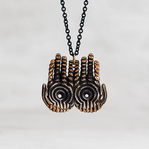 A pair of hands pendant necklace with a textured bronze finish and black accents, suspended from a black chain against a light gray background.