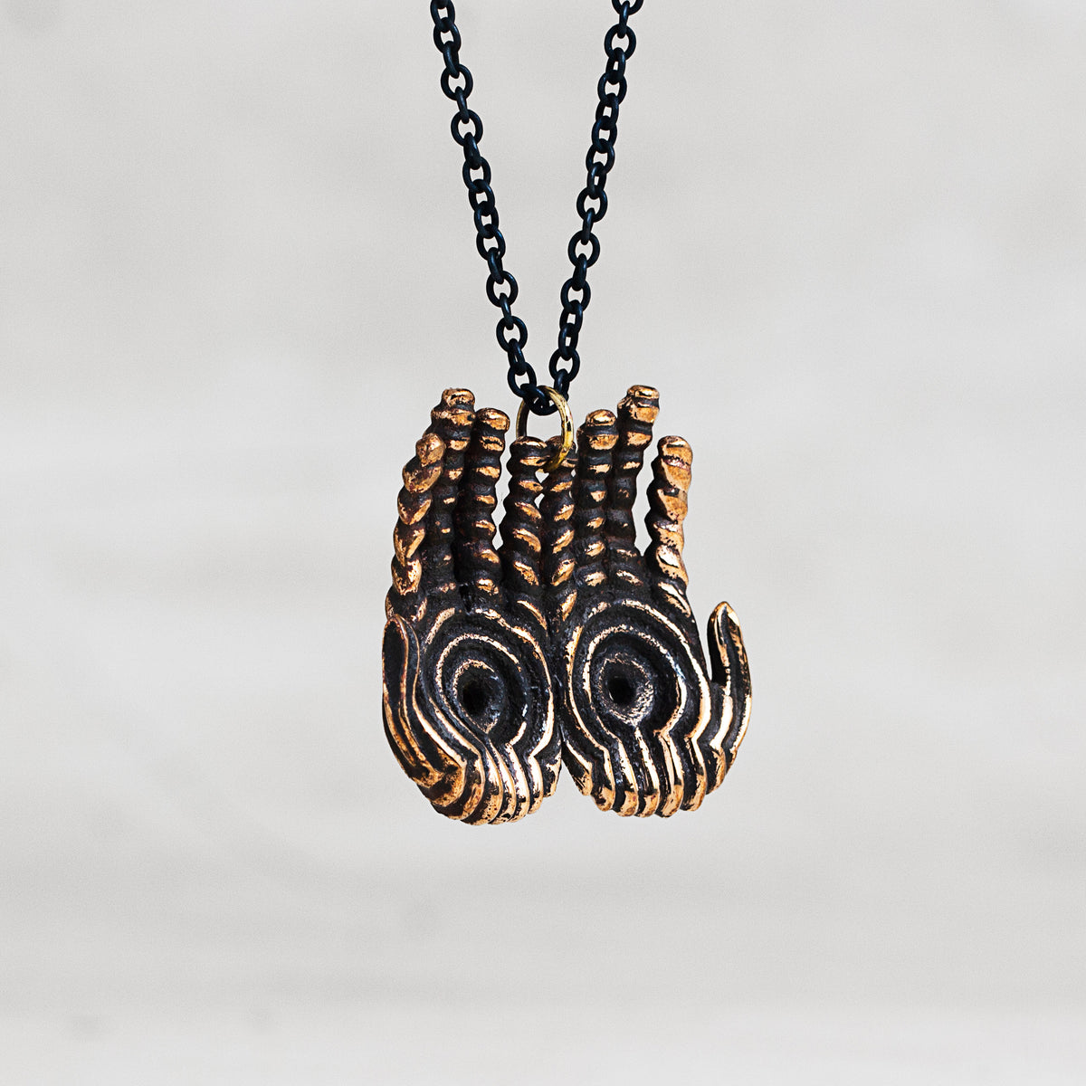 A close-up of a necklace featuring two sculpted hand pendants with black and gold striped patterns, suspended from a black chain against a light gray background.