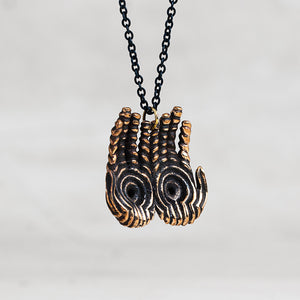 A close-up of a necklace featuring two sculpted hand pendants with black and gold striped patterns, suspended from a black chain against a light gray background.