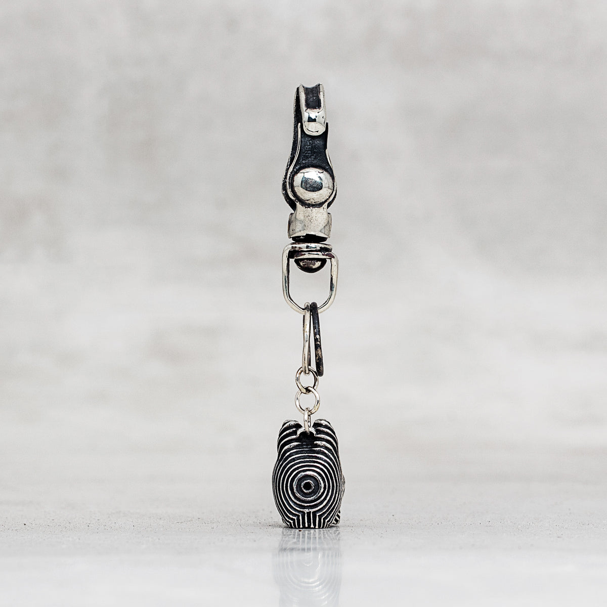 A silver-toned metal carabiner clip with a dangling charm shaped like a black-and-white spiral-patterned head, shown against a light gray textured background.