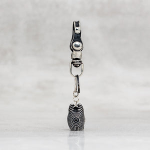 A silver-toned metal carabiner clip with a dangling charm shaped like a black-and-white spiral-patterned head, shown against a light gray textured background.