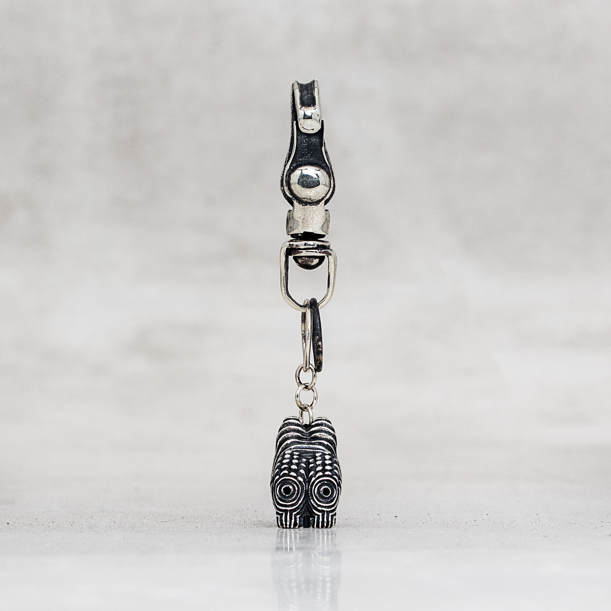 A silver-toned metal clasp with a dangling charm featuring black oxidized detailing, shown against a light gray textured background.