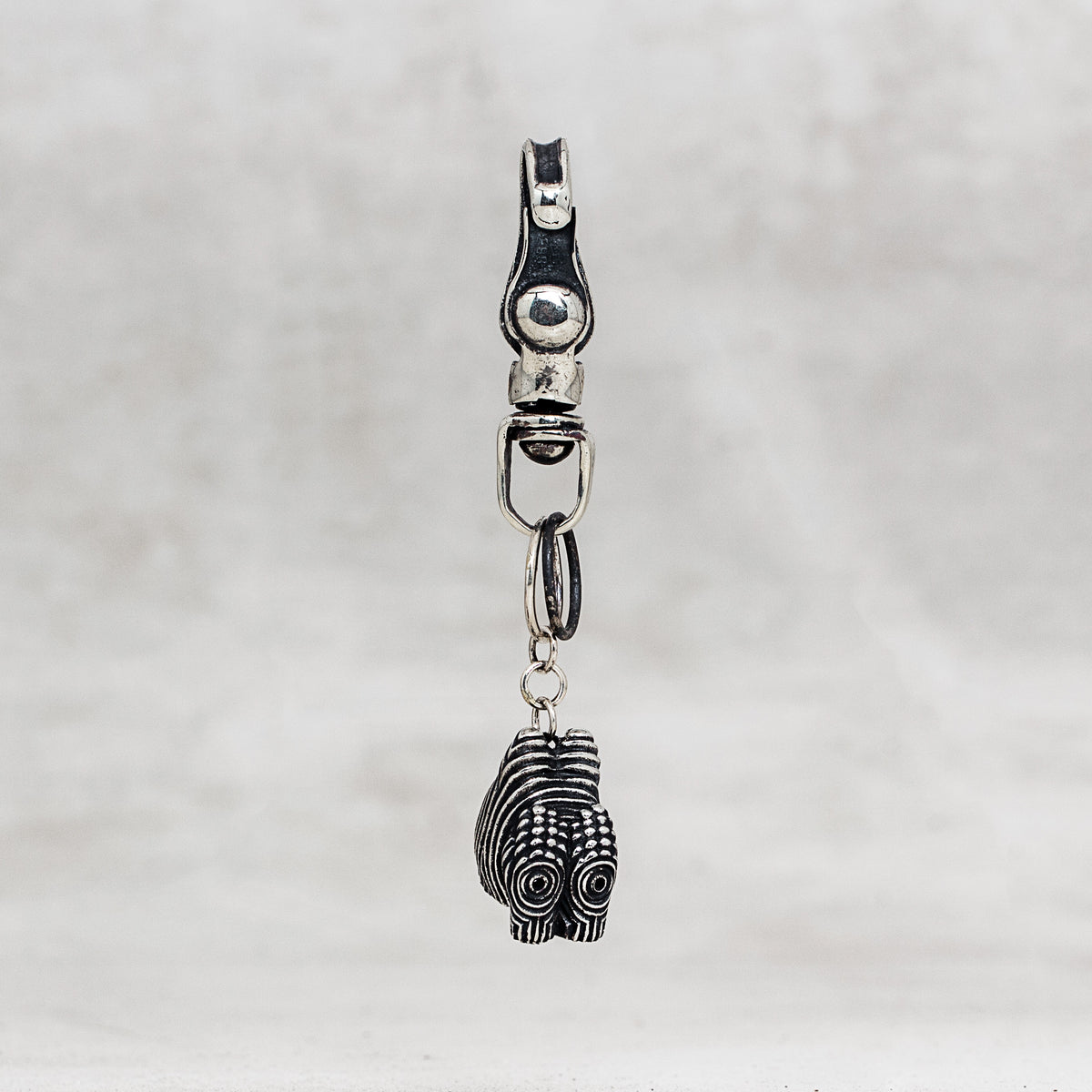 A silver-toned metal keychain or bag charm featuring a small carved pendant with black oxidized detailing, shown against a light gray textured background.